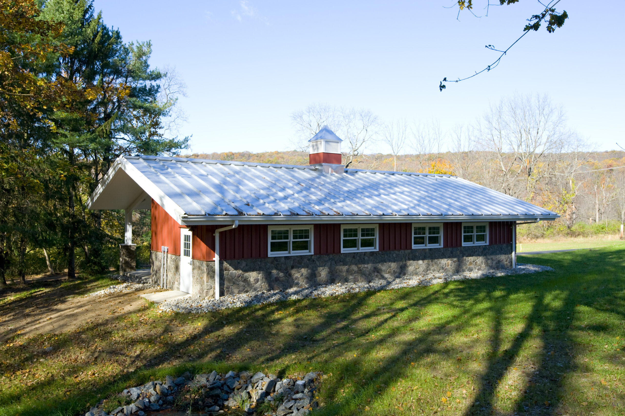 Exterior of a small red metal building with a standing seam metal roof, stone base, and cupola, set on a grassy, wooded site.
